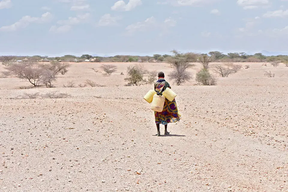 A women carries three large empty water jugs over arid land